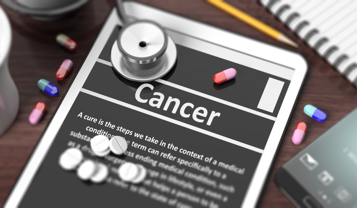 Tablet with "Cancer" on screen, stethoscope, pills and objects on wooden desktop.